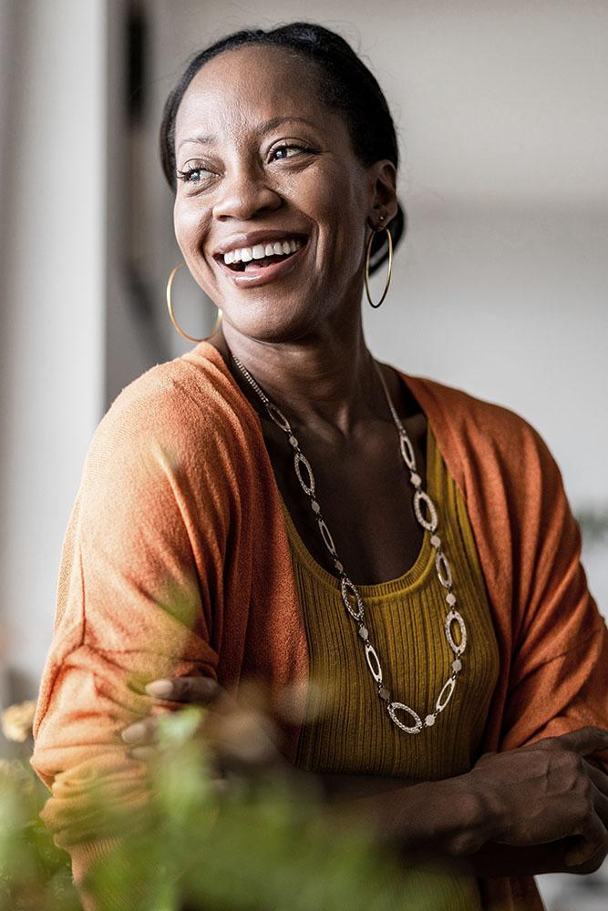 A smiling woman with hoop earrings and a long necklace, wearing an orange cardigan over a mustard yellow top, stands with her arms crossed in a softly lit indoor setting.