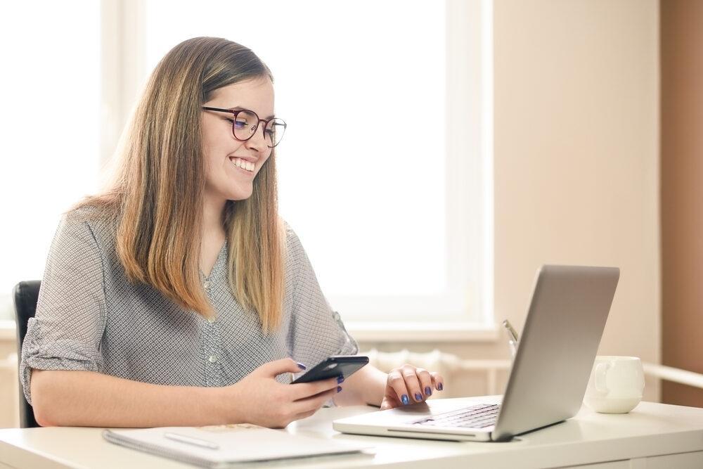 A smiling woman with glasses sits at a desk, holding a smartphone and looking at a laptop. A notebook and cup are on the desk, and soft daylight enters through a window behind her.