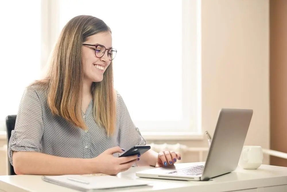 A smiling woman with glasses sits at a desk, holding a smartphone and looking at a laptop. A notebook and cup are on the desk, and soft daylight enters through a window behind her.
